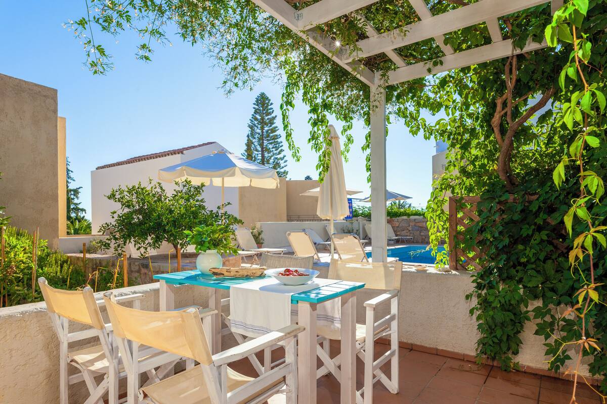 Un patio al aire libre con mobiliario blanco, una piscina y una pérgola con hiedra colgante.