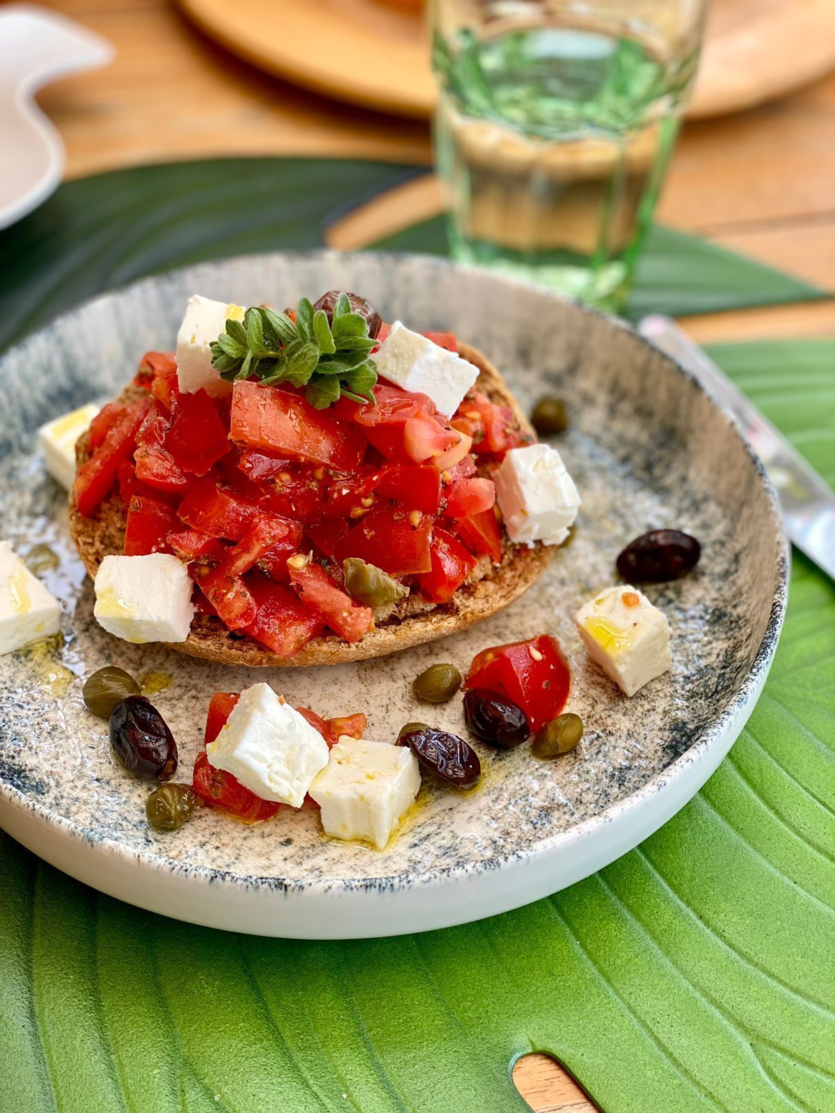 Un plato con una rebanada de pan cubierta de tomates, aceitunas y queso feta, decorada con perejil.