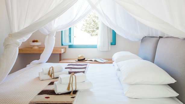 Une chambre à coucher avec un lit à baldaquin, une table de chevet sur laquelle se trouvent un chapeau et un livre, et une fenêtre avec des volets bleus.