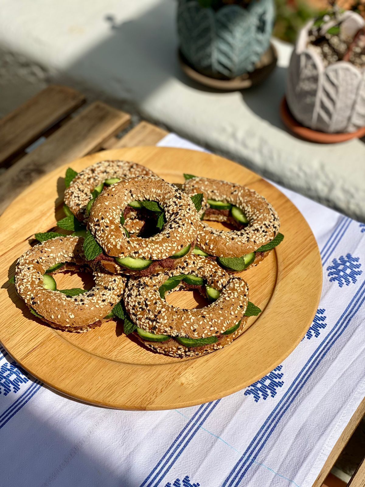 A wooden plate with bagels topped with sesame seeds and cucumber slices, placed on a table with a white cloth and blue patterns.