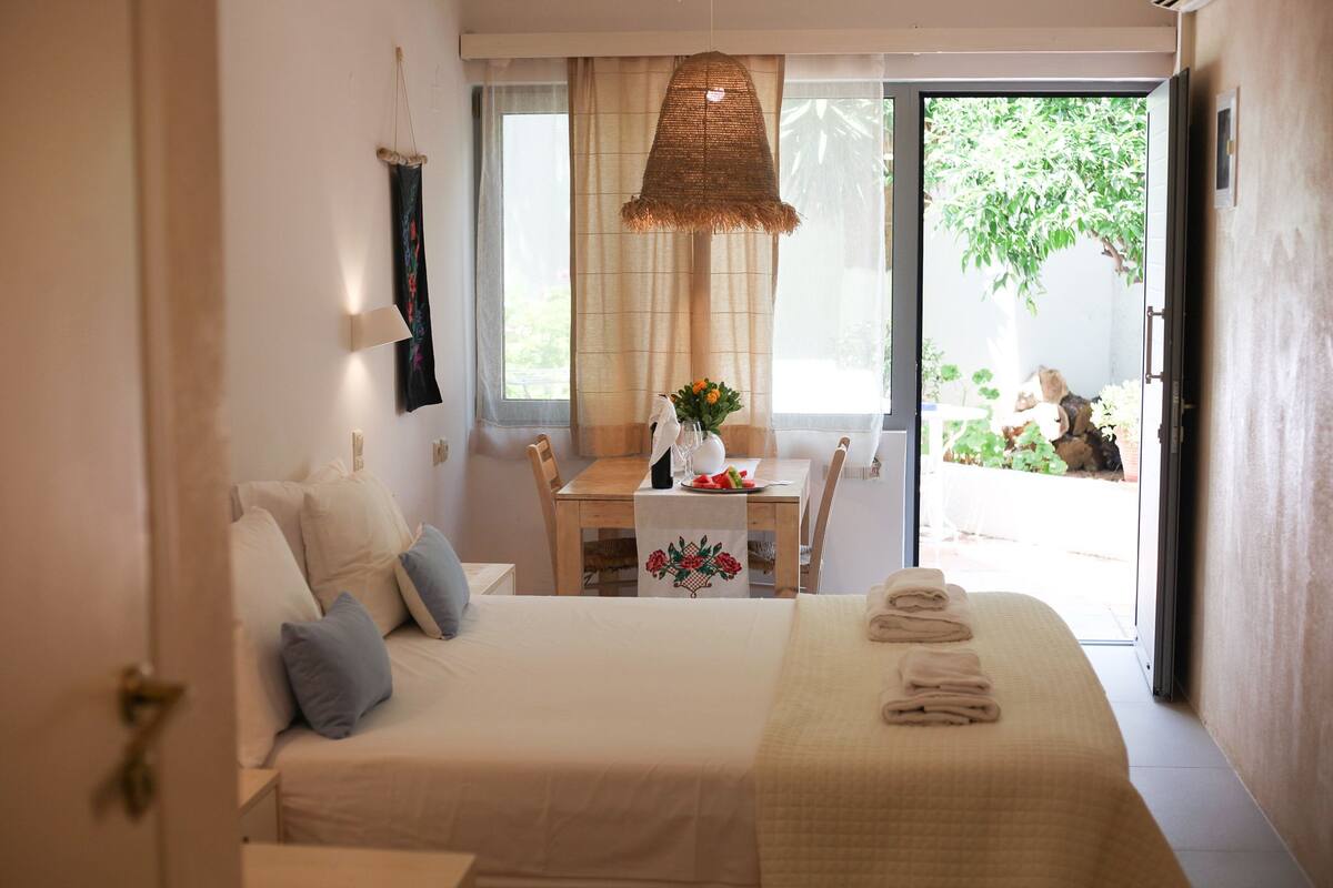 Un dormitorio con una cama, una mesa con un florero y fruta, y vistas al exterior.