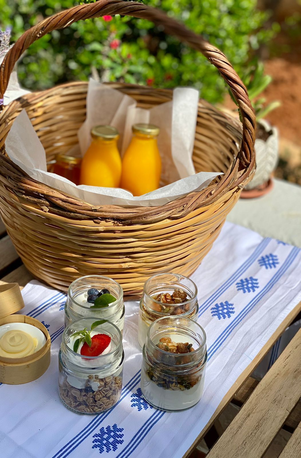 Un panier en osier contenant deux bouteilles de jus d’orange, un bol en bois et trois bocaux en verre de différentes sortes, le tout posé sur une nappe à motifs bleus.