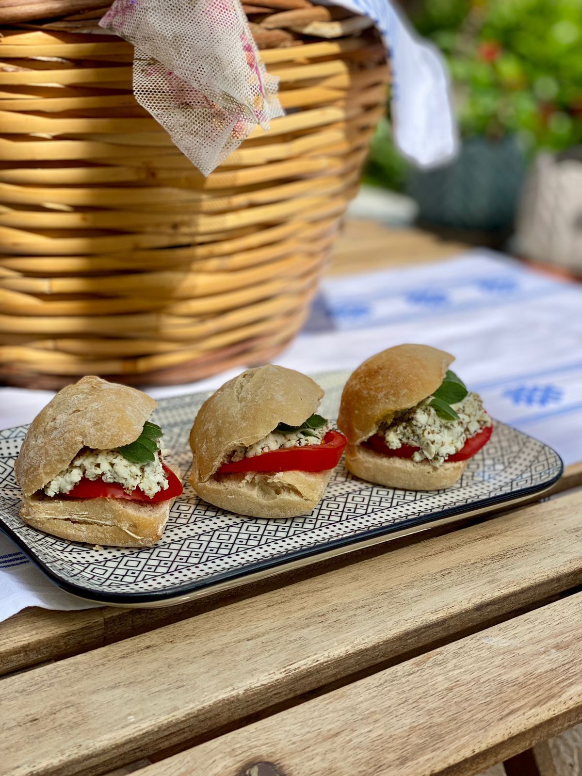 Three sandwiches on a patterned plate on a wooden table with a woven basket in the background.