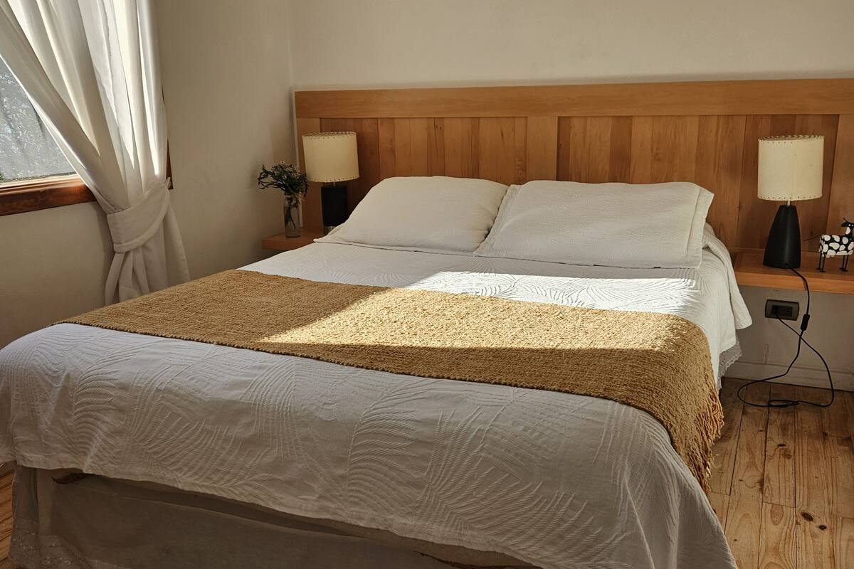 A bedroom with a wooden headboard, two bedside tables with lamps, a bed with white linens, and a window with curtains.