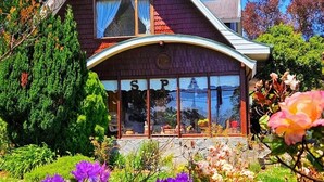 A two-story house with a prominent front porch, large windows, and a garden with various flowers. - Canales del Sur (Puerto Varas)