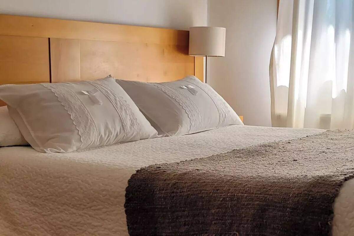 A neatly made bed with a white duvet and two pillows, a wooden headboard, and a bedside lamp.