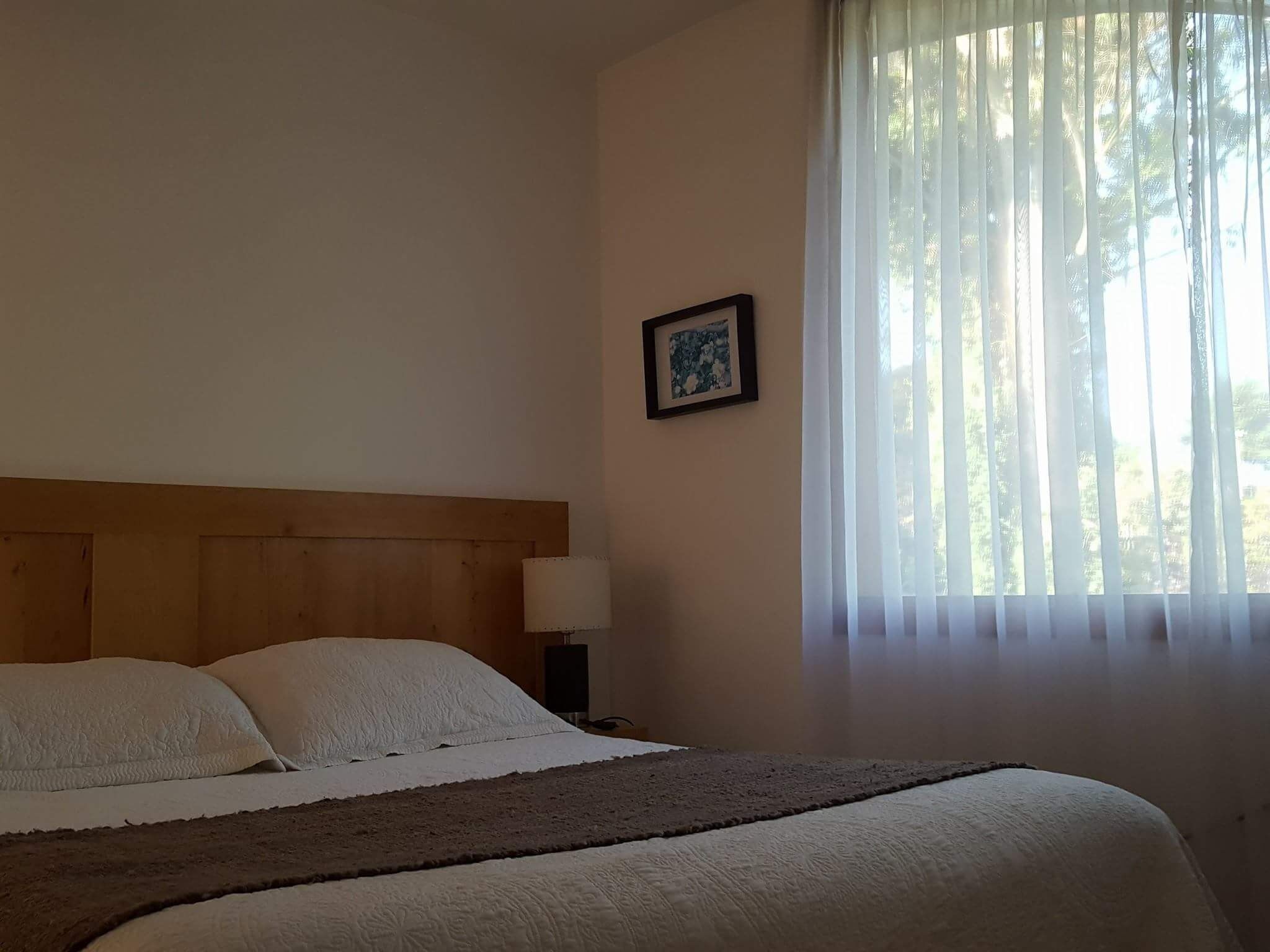 Un dormitorio con cabecera de madera, cama con ropa de cama blanca, mesita de noche con lámpara y ventana con cortinas traslúcidas.