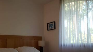 Un dormitorio con cabecera de madera, cama con ropa de cama blanca, mesita de noche con lámpara y ventana con cortinas traslúcidas.