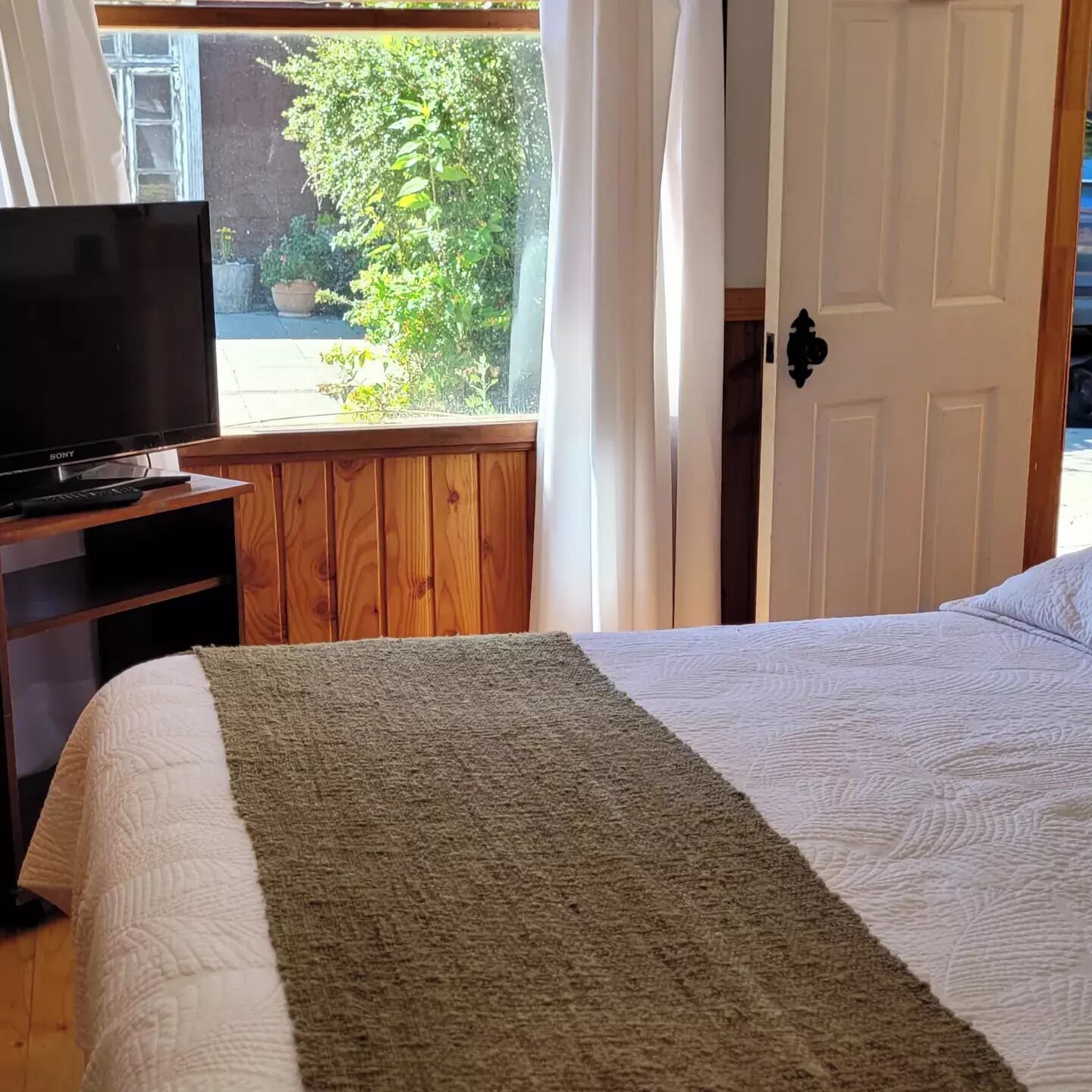 A bedroom with a bed, a television on a wooden stand, a window with curtains, and a door with a handle.