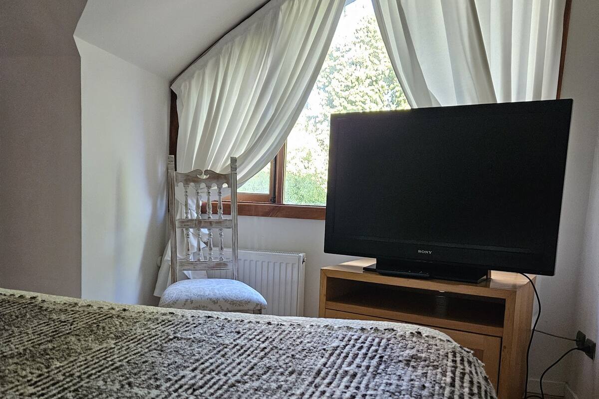 A bedroom with a large bed, a television on a wooden stand, a radiator, and a window with curtains.
