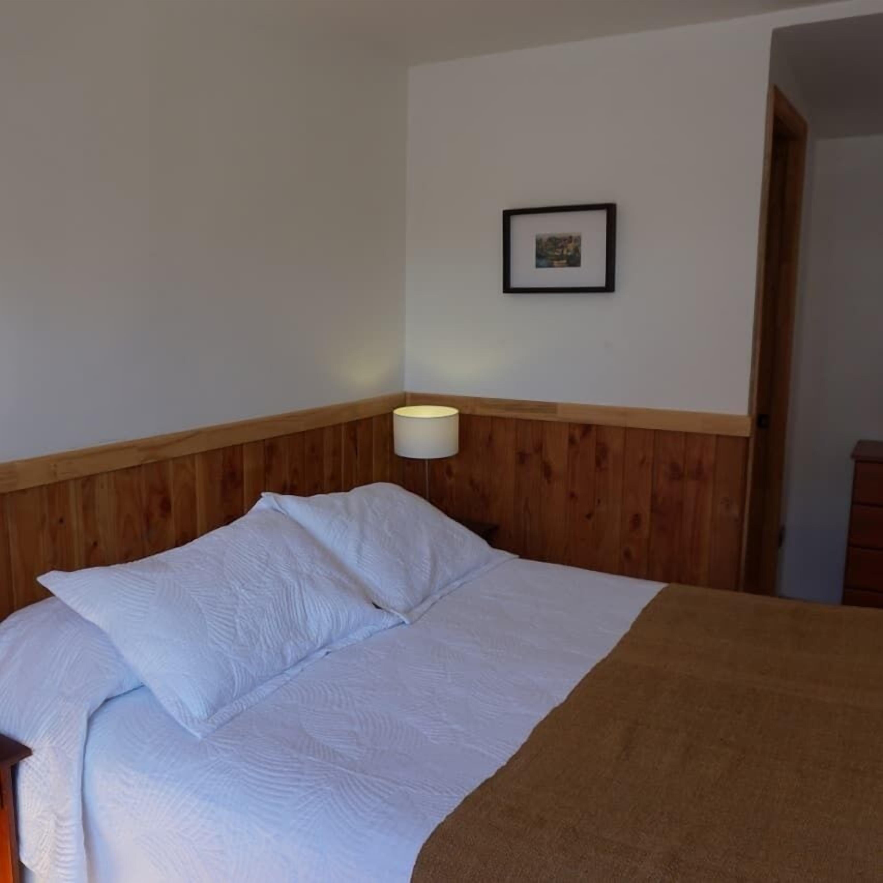 A neatly made bed with white linens and a brown throw blanket, a bedside lamp, and a framed picture on the wall.