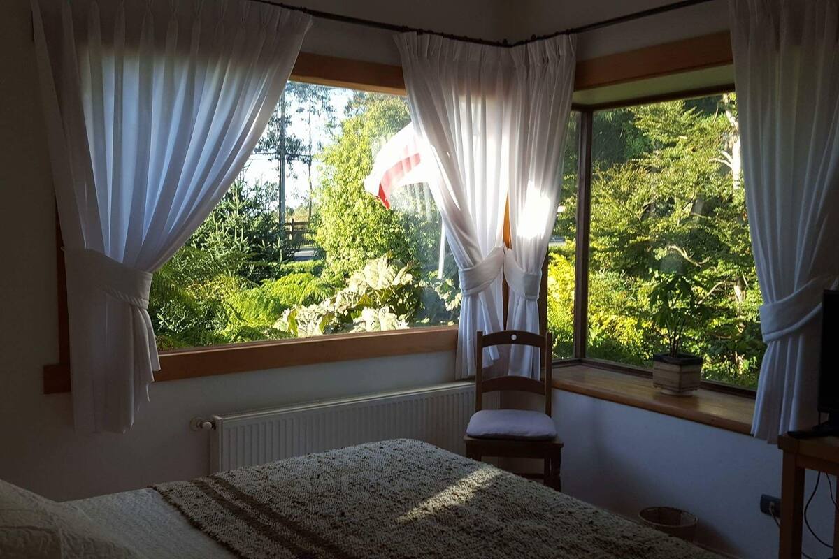 A bedroom with a bed, a chair, a window with curtains, and a view of greenery outside.