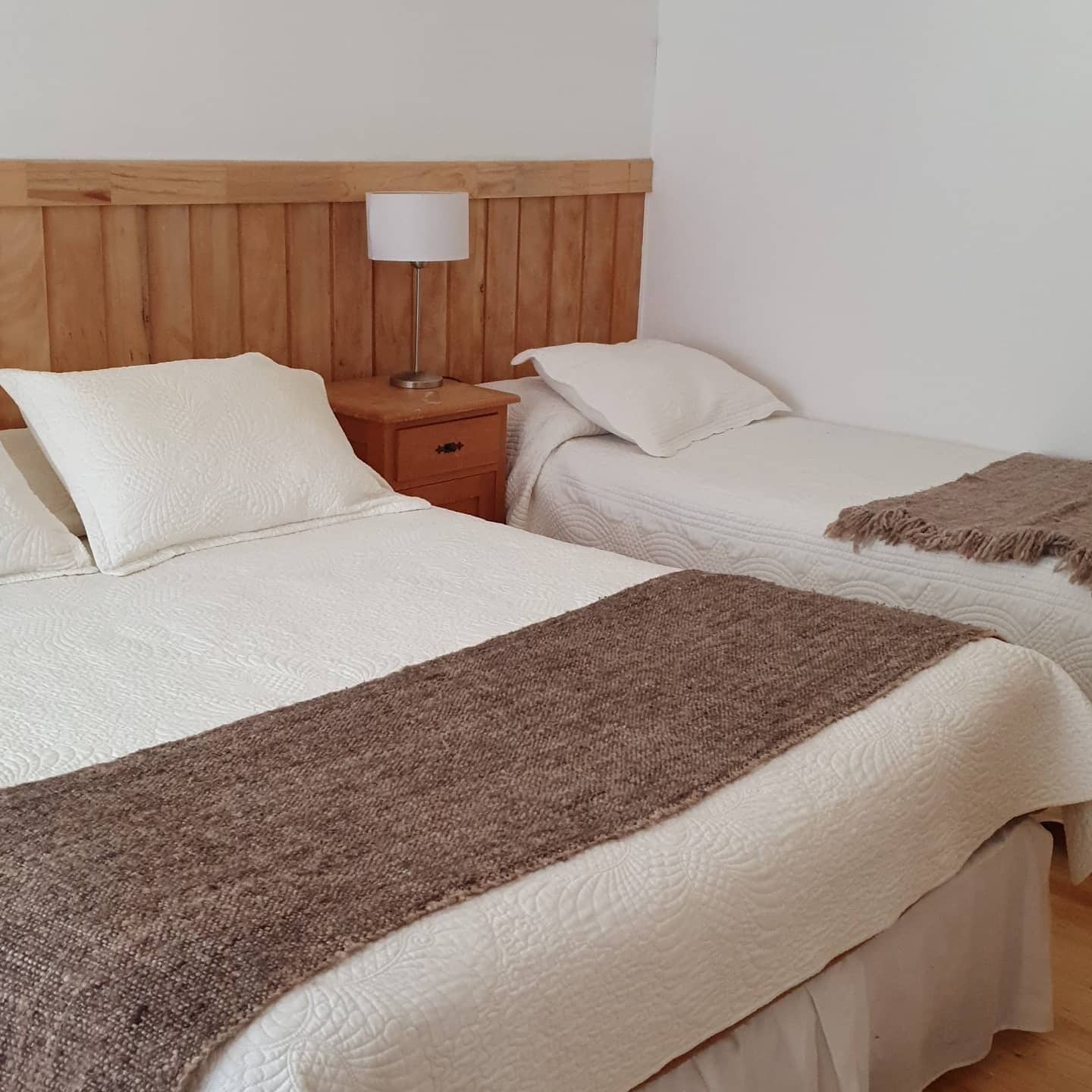 Une chambre d’hôtel avec deux lits, une tête de lit en bois, une table de chevet avec une lampe et un plaid marron.