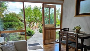A room with a wooden floor, a sofa, a dining table with chairs, and a sliding glass door leading to an outdoor area with greenery. - Canales del Sur (Puerto Varas)
