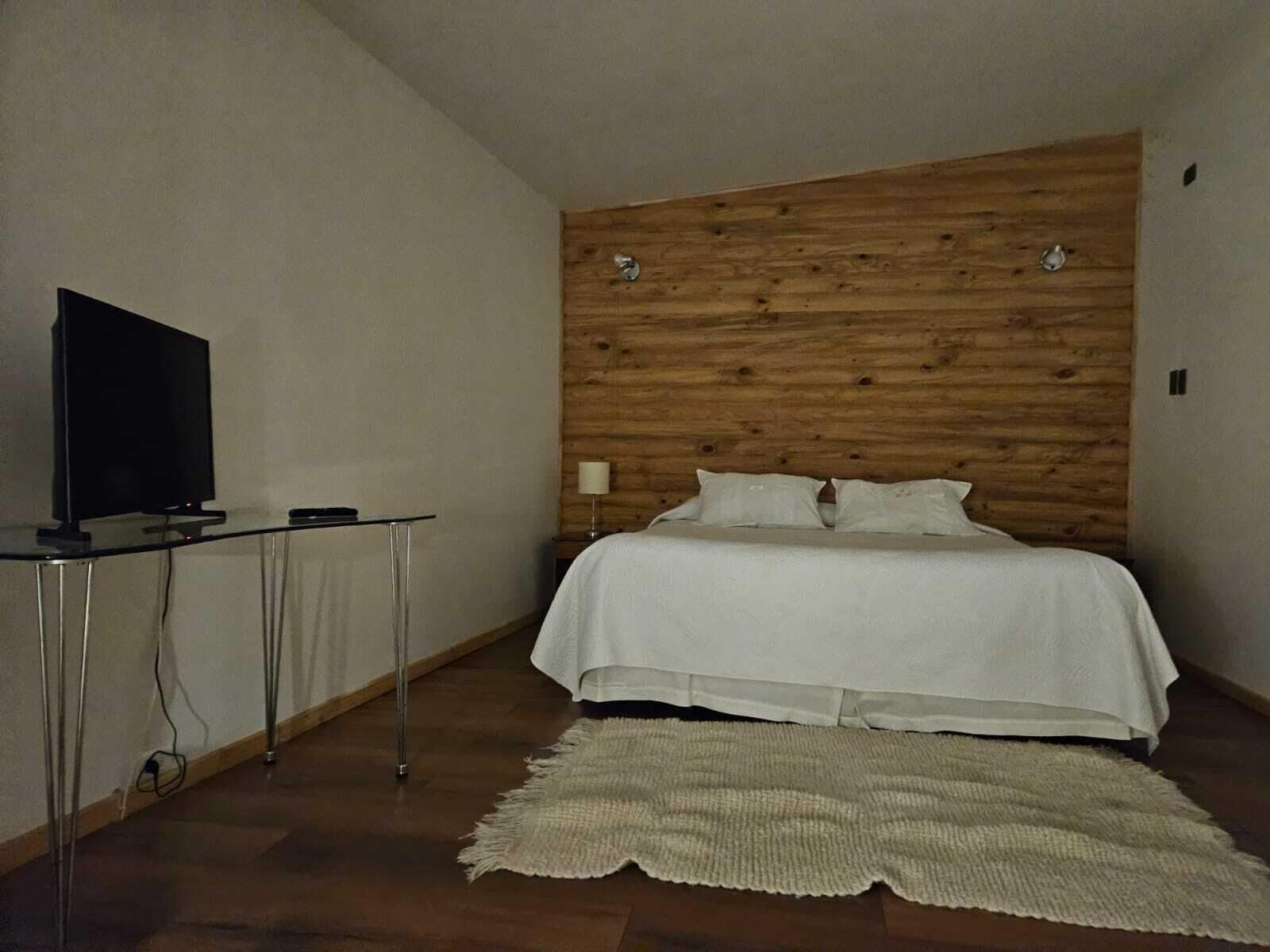 A bedroom with a wooden headboard, a bed with white bedding, a television on a glass table, and a woven rug on the floor.