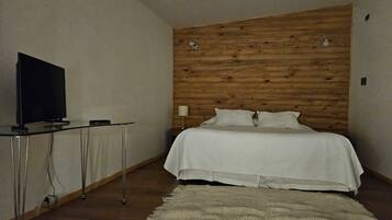 Un dormitorio con cabecera de madera, cama con ropa blanca, televisor sobre una mesita de vidrio y alfombra tejida en el suelo.