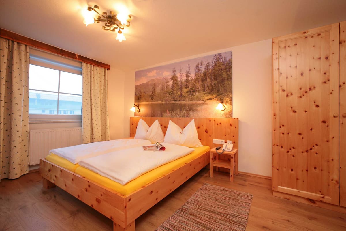 A wooden bed with a yellow mattress, a bedside table with a phone, and a painting of a forest on the wall.