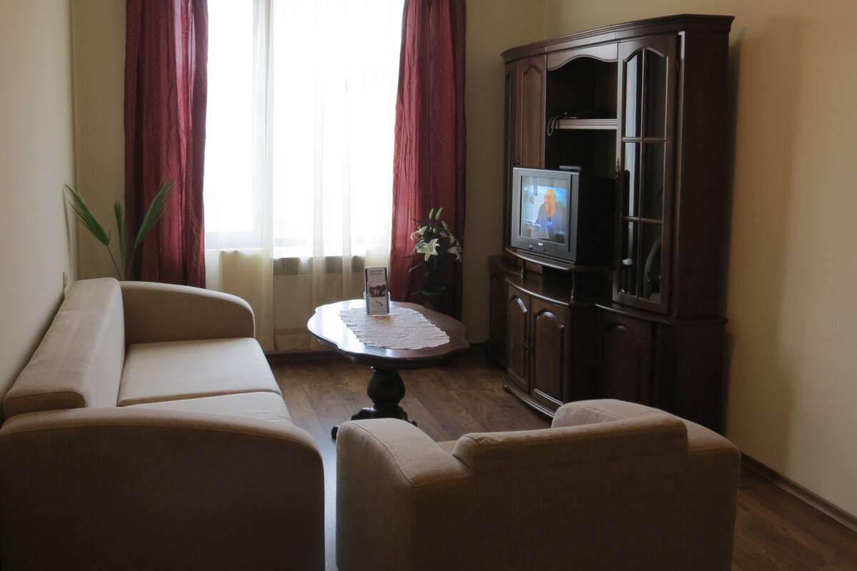 A living room with a sofa, a small table with a placemat, a television on a cabinet, and a window with curtains.