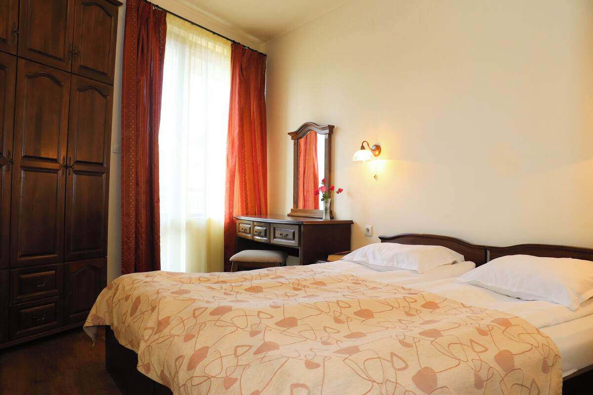 A hotel room with a large wardrobe, a bed with patterned bedding, a desk with a mirror, and a wall-mounted light fixture.