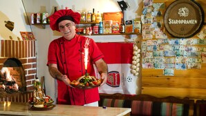 Een kok in een rood uniform houdt een dienblad met eten vast, met een bakoven en een muur met diverse voorwerpen en geldnotulen.