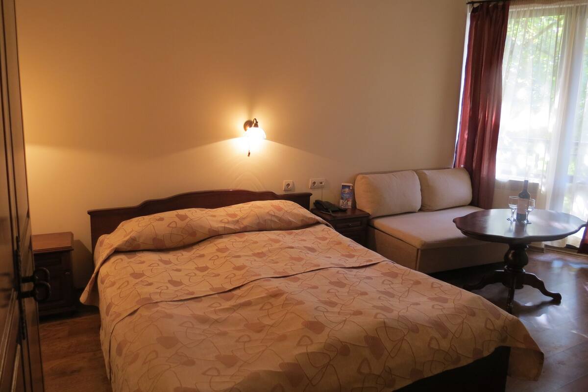 A hotel room with a bed, a sofa, a small table with a bottle and glasses, a wall-mounted light, and a window with curtains.