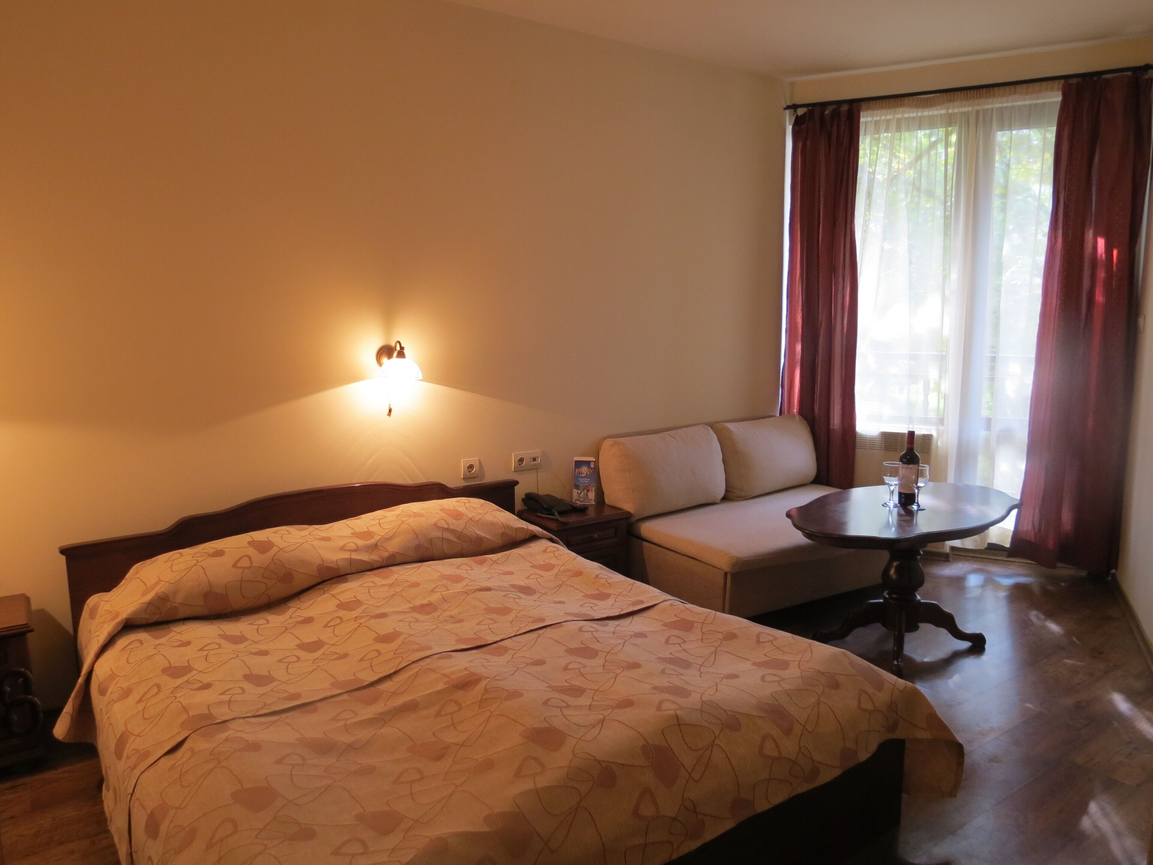 A hotel room with a bed, a sofa, a small table with a wine bottle and glasses, and a wall-mounted lamp.