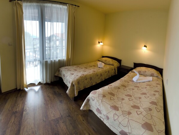 A hotel room with two beds, a window with curtains, a wooden floor, and wall-mounted lights. - Apartments & Studios Mountain Romance (Bansko)