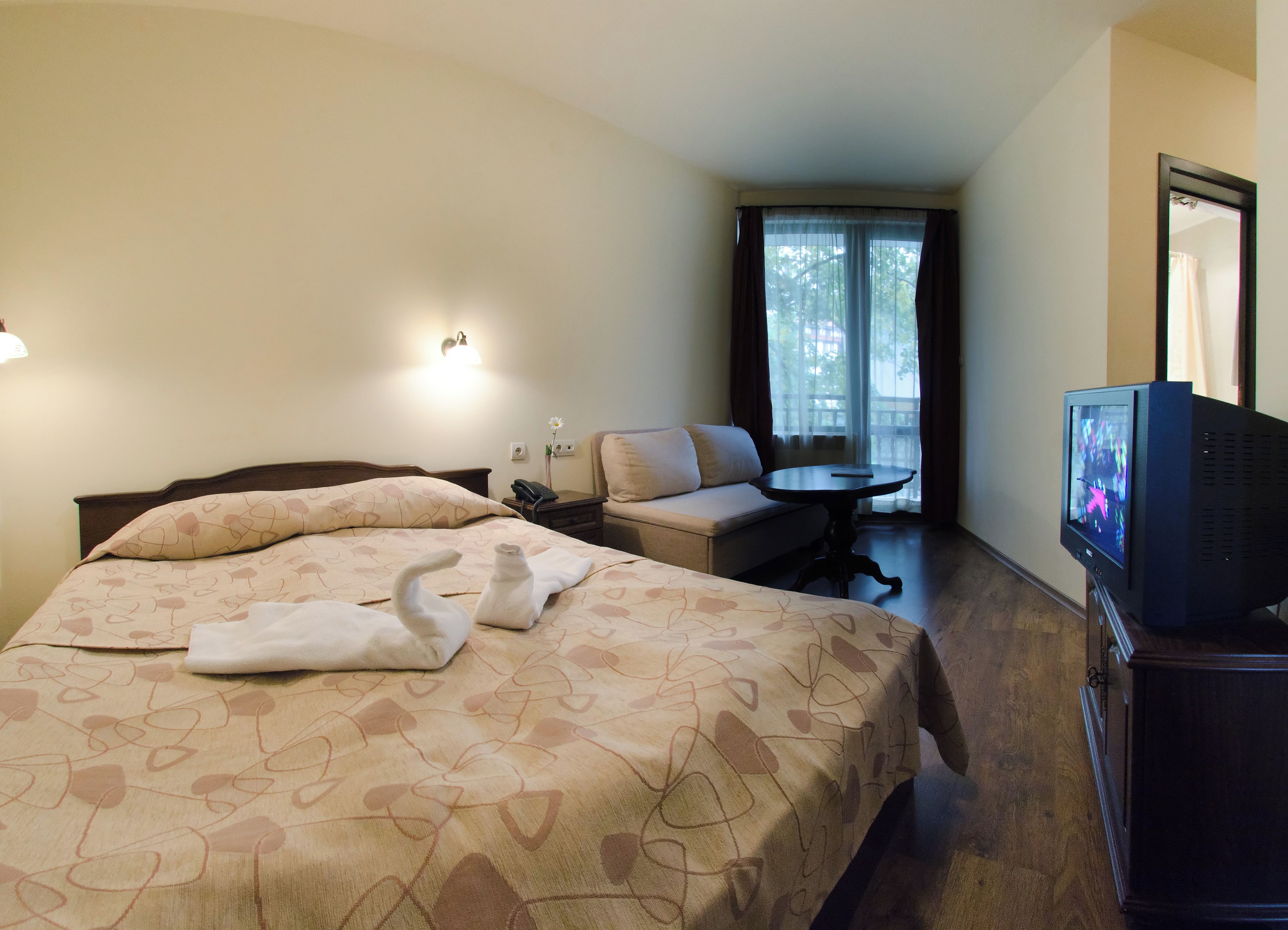 Una habitación de hotel con cama, sofá, una mesita, televisor y ventana con cortinas.