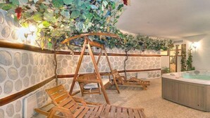 Een wellnessruimte met een stenen muur, houten ligbedden, een jacuzzi en een hangstoel aan de muur.