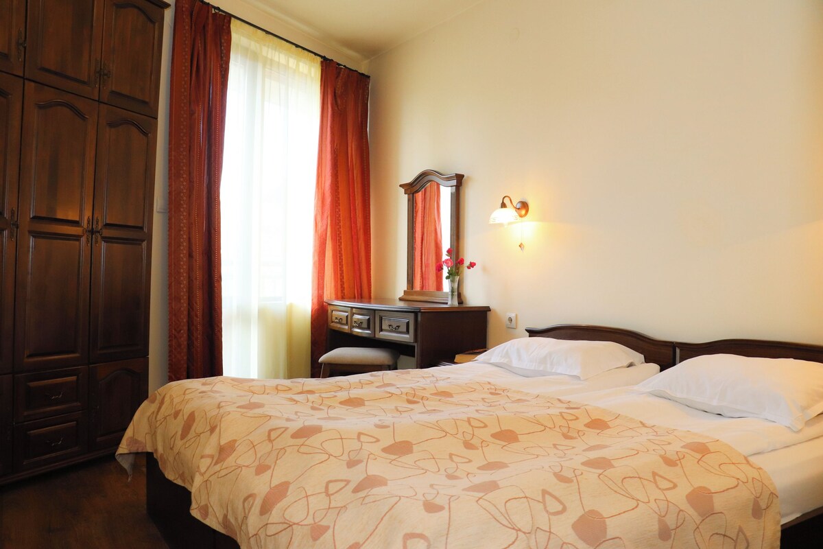 Una habitación de hotel con un armario grande, una cama con ropa de cama estampada, un escritorio con espejo y una lámpara fijada a la pared.