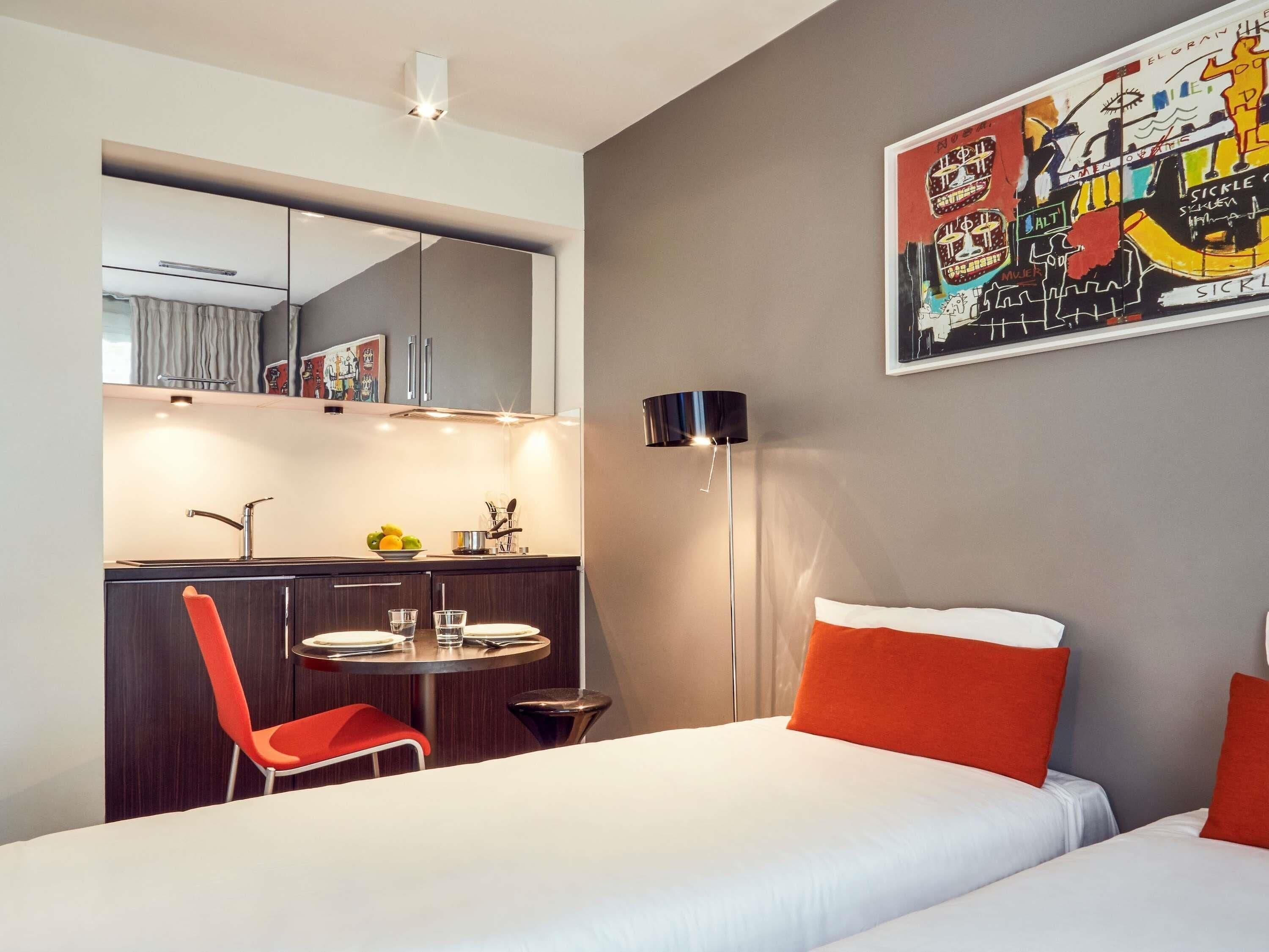Una habitación de hotel con una cama, un escritorio con silla, un espejo, una lámpara de pie y un cuadro de colores en la pared.