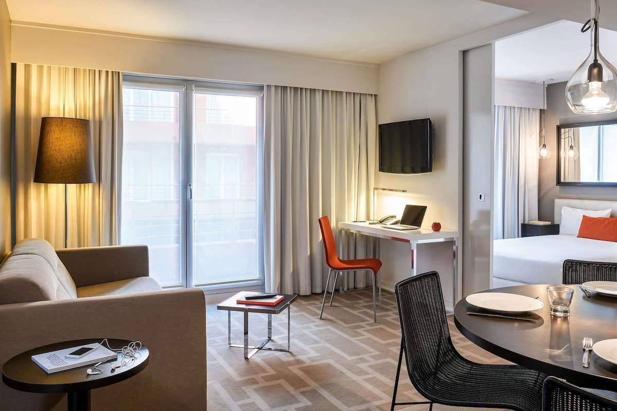 Ein Hotelzimmer mit einer Sitzecke, einem Schreibtisch mit Laptop, einem Fernseher, einem Bett und einem Essbereich mit Stühlen und Tisch.