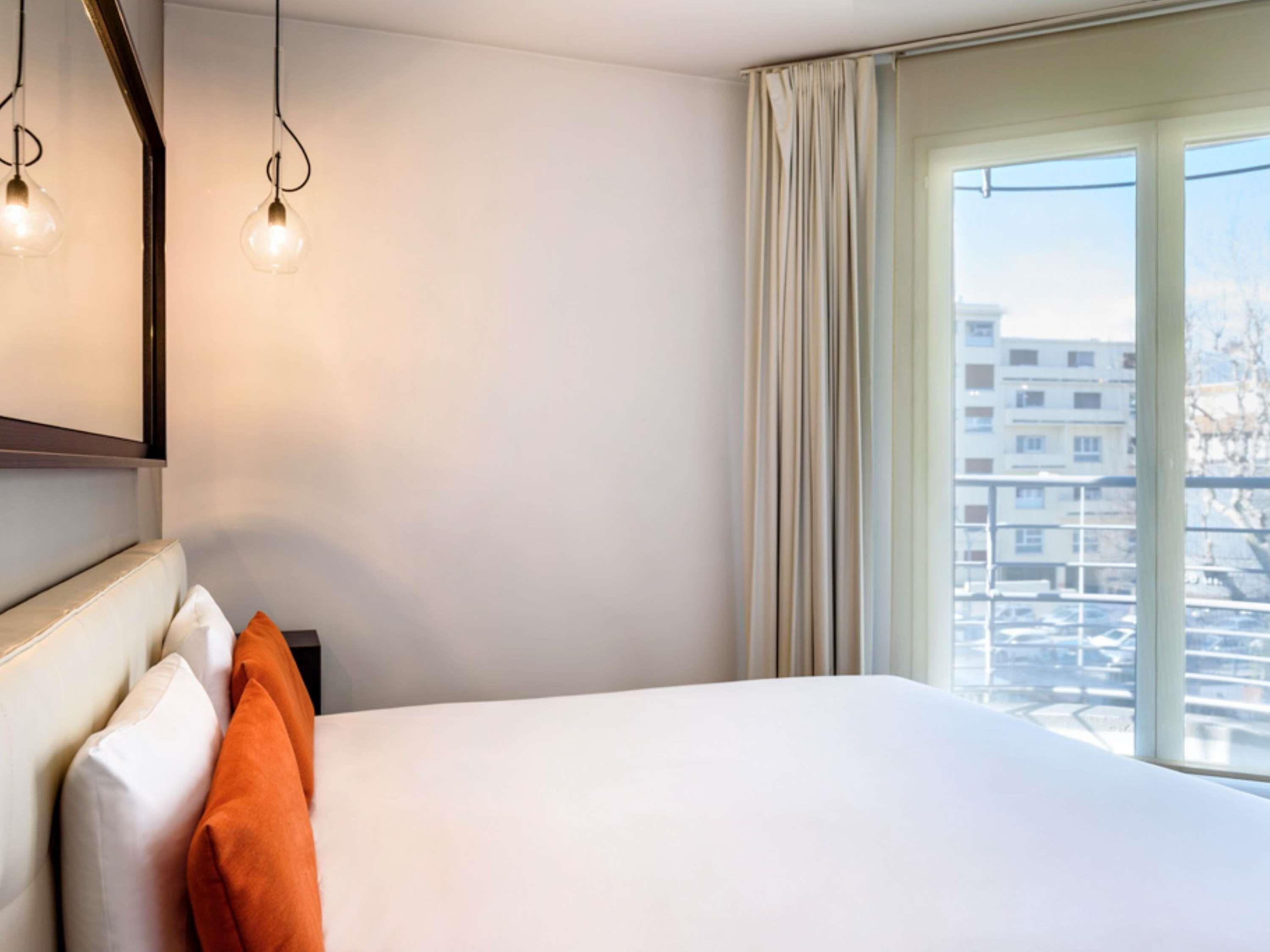 A hotel room with a bed, a nightstand, a mirror, and a window with a view of buildings.