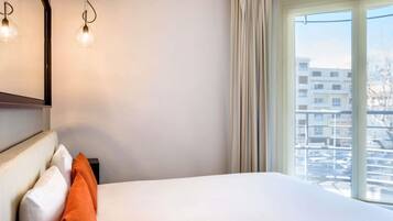 Una habitación de hotel con cama, mesita de noche, espejo y ventana con vistas a edificios.