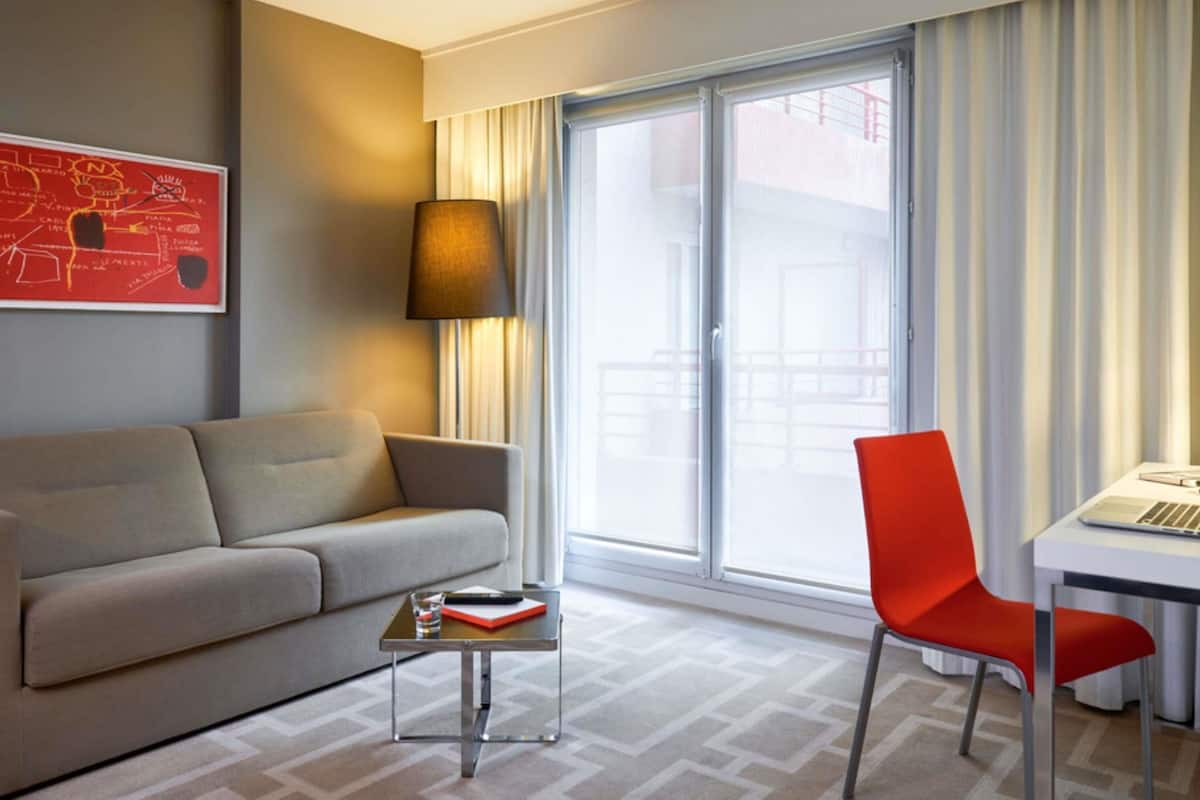 Ein Hotelzimmer mit einer Couch, einem kleinen Tisch mit Fernbedienung, einem roten Sessel, einem Schreibtisch mit Laptop und einem großen Fenster mit Vorhängen.