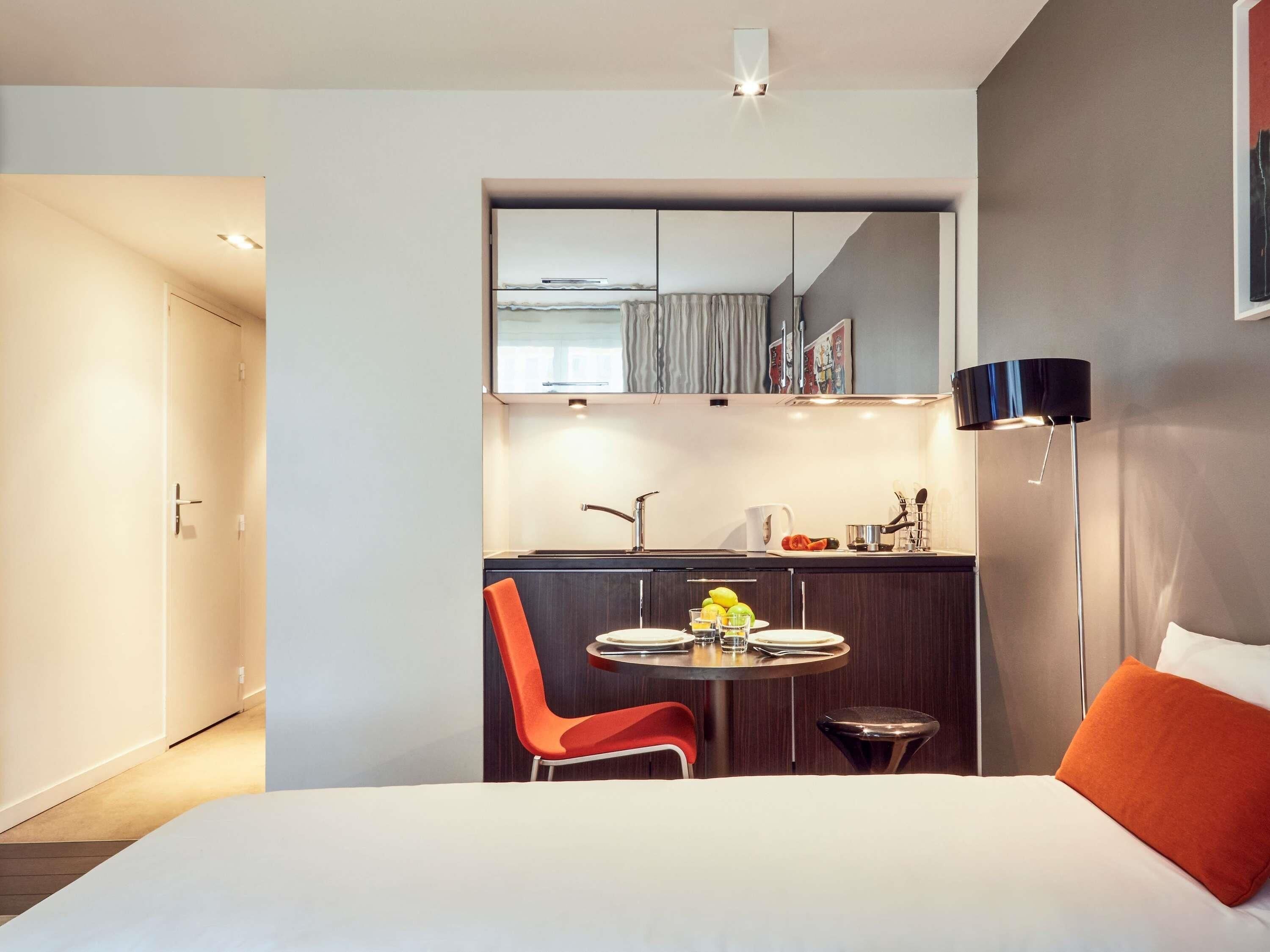 Una habitación de hotel moderna con una cama, una mesa de comedor con sillas, una pequeña cocina y una lámpara de noche.