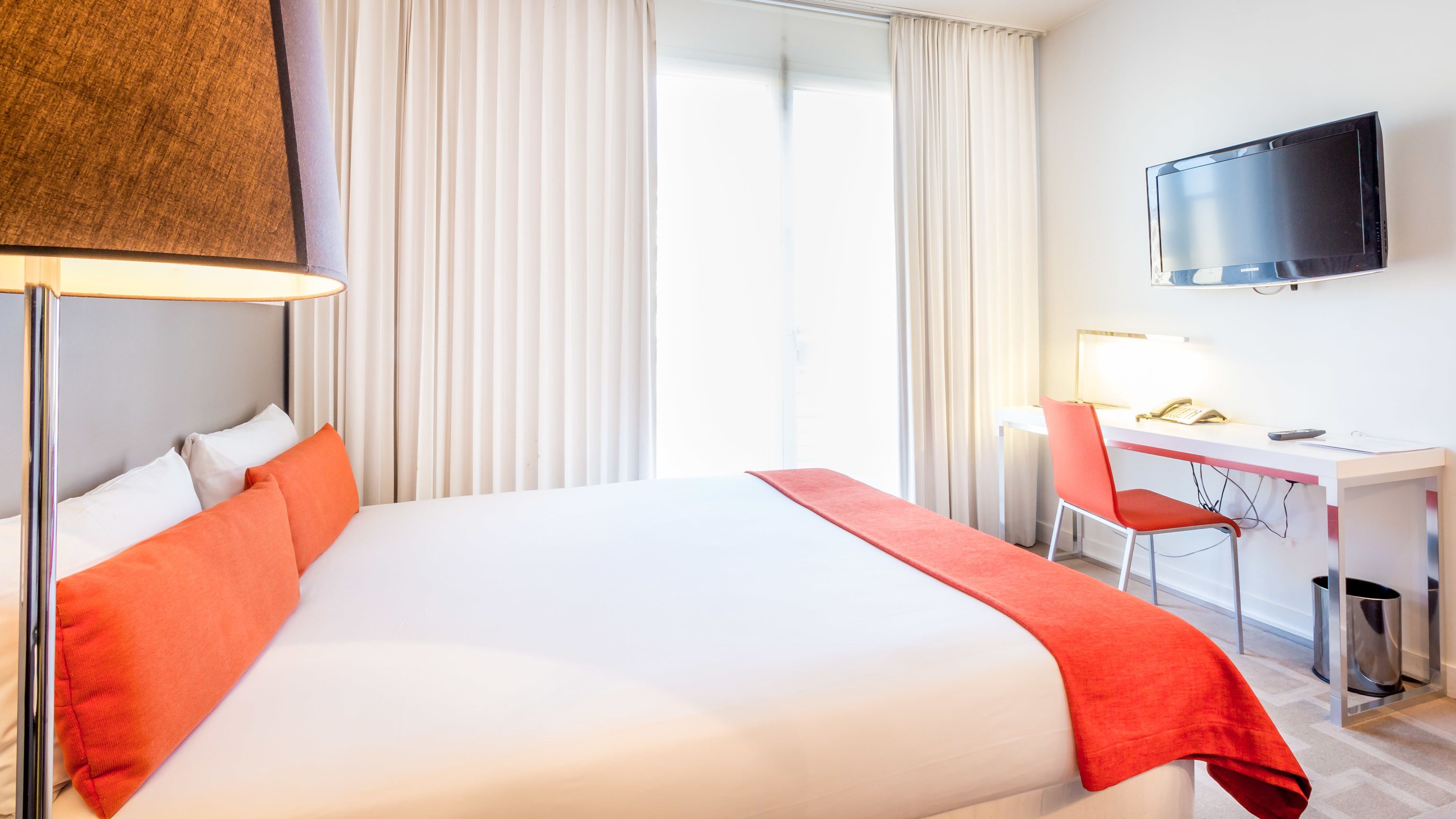 Quarto de hotel com cama, escrivaninha, cadeira, televisão e janela com cortinas.