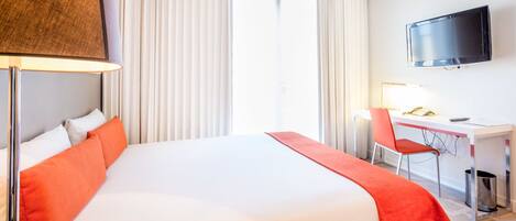 Quarto de hotel com cama, escrivaninha, cadeira, televisão e janela com cortinas.