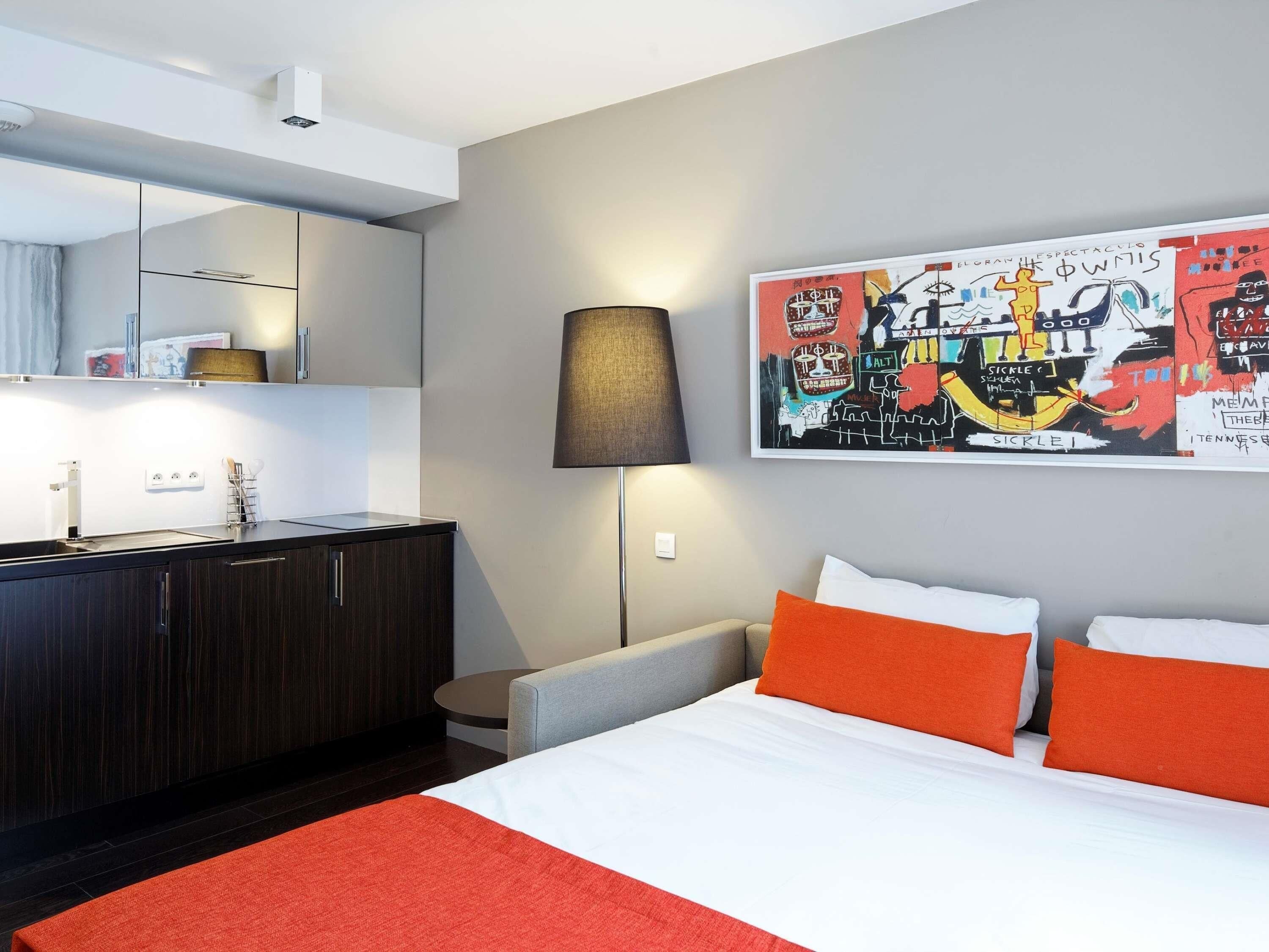 Et moderne hotelværelse med seng, skrivebord, stol, lampe og et stort abstrakt maleri på væggen.