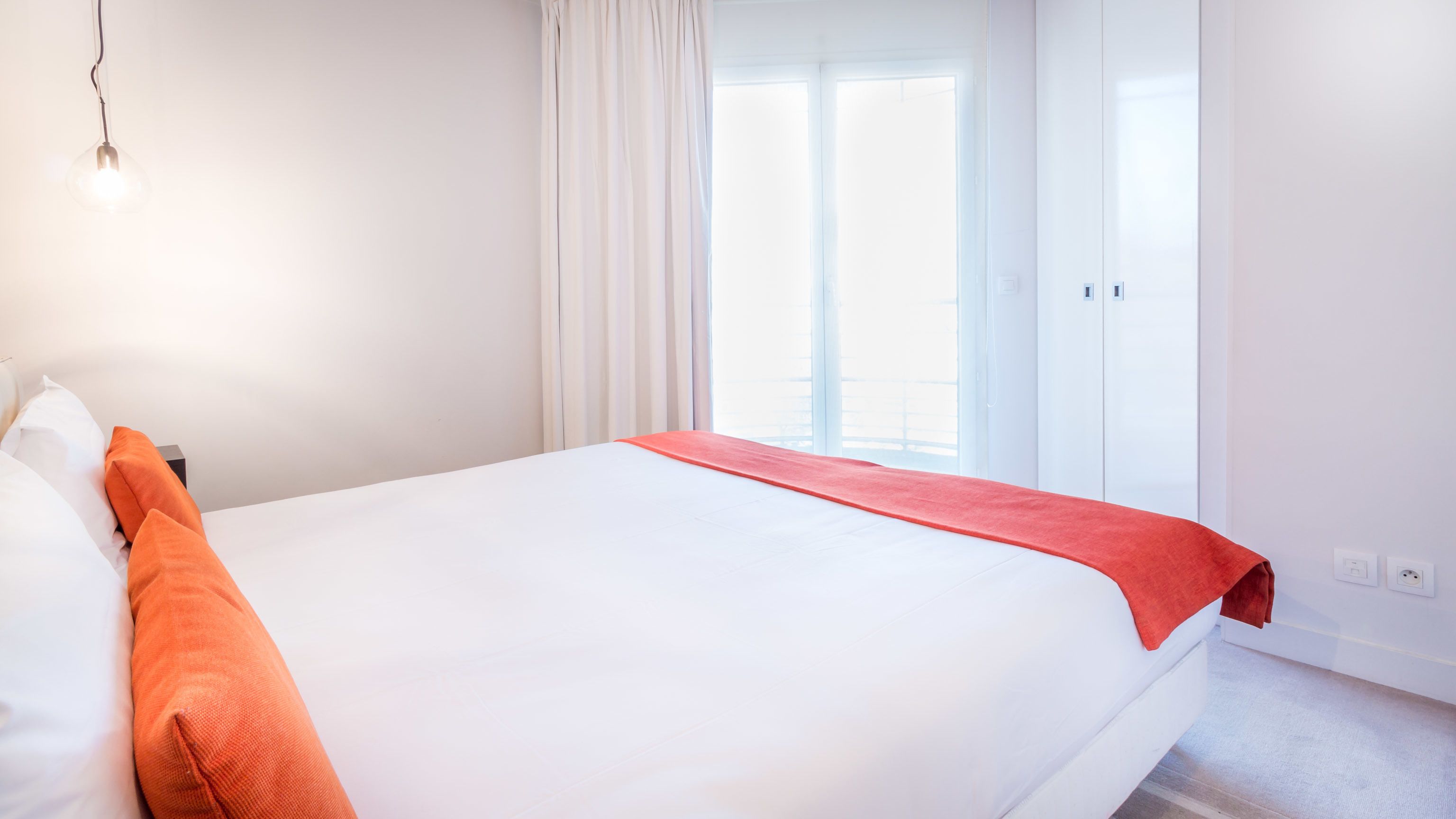 Een hotelkamer met een groot bed, witte muren, een raam met gordijnen en een rode plaid.