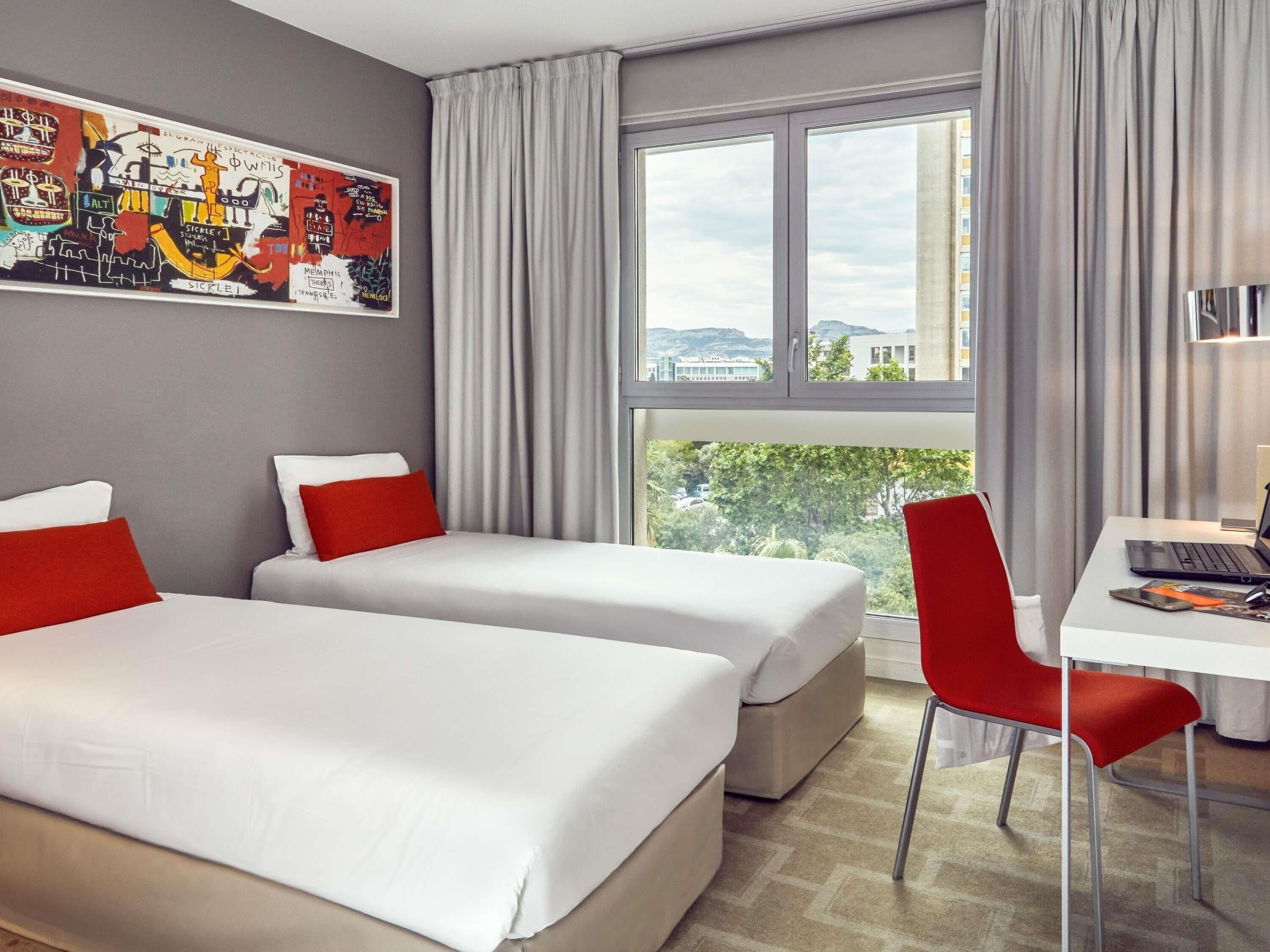 A hotel room with two beds, a desk, a chair, a large window, and a cityscape view.