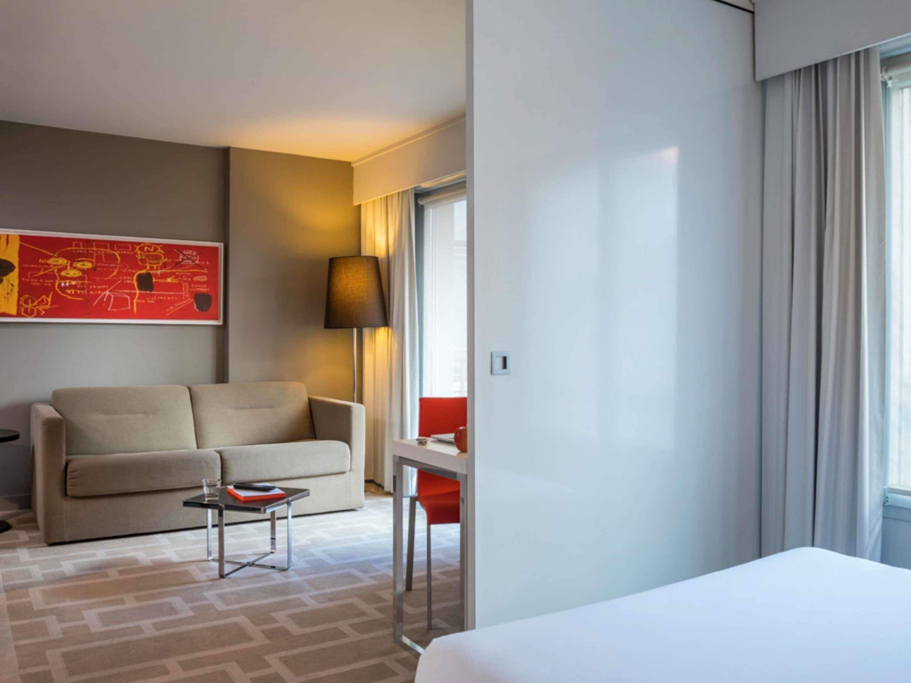 Une chambre d’hôtel avec un lit, un canapé, une petite table, une lampe, une fenêtre avec des rideaux et un mur d’accent rouge.