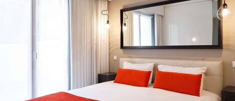 Habitación de hotel con una cama grande, dos almohadas, una manta roja, un espejo grande y una ventana con cortinas.