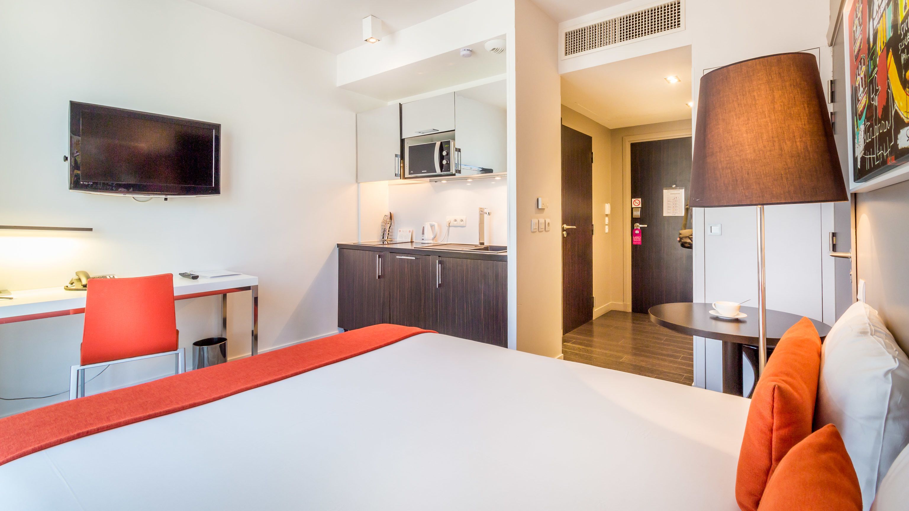 A hotel room with a bed, a desk, a chair, a TV, a microwave, and a door leading to a bathroom.