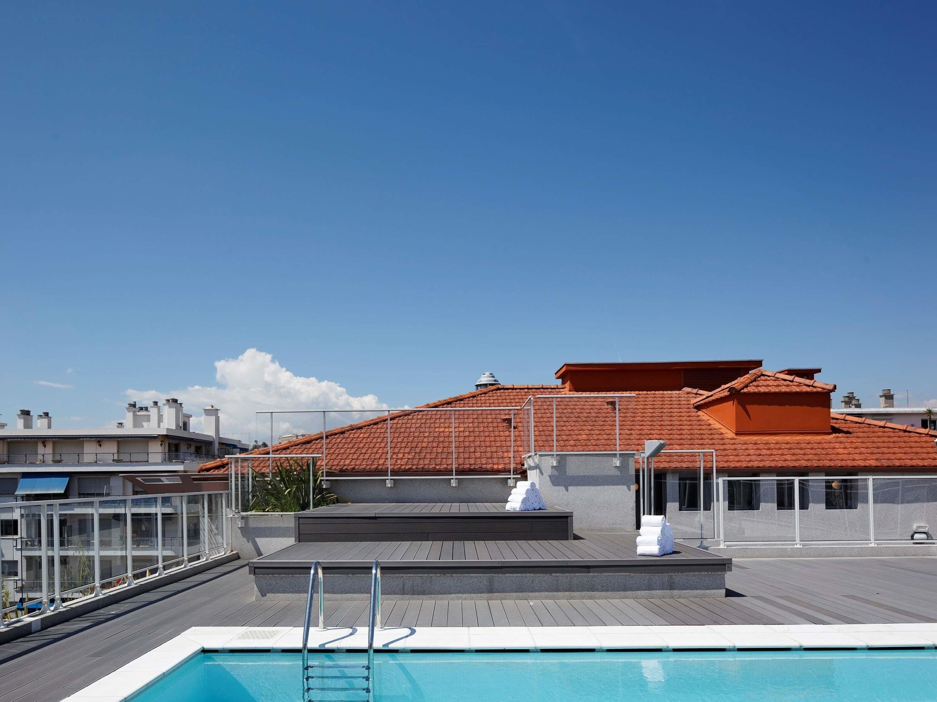 A rooftop swimming pool with a clear view of the sky and a building with a red-tiled roof in the background.