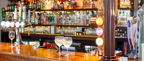 Uma área de bar com balcão de madeira, vários dispensadores de bebidas iluminados e uma variedade de garrafas ao fundo.