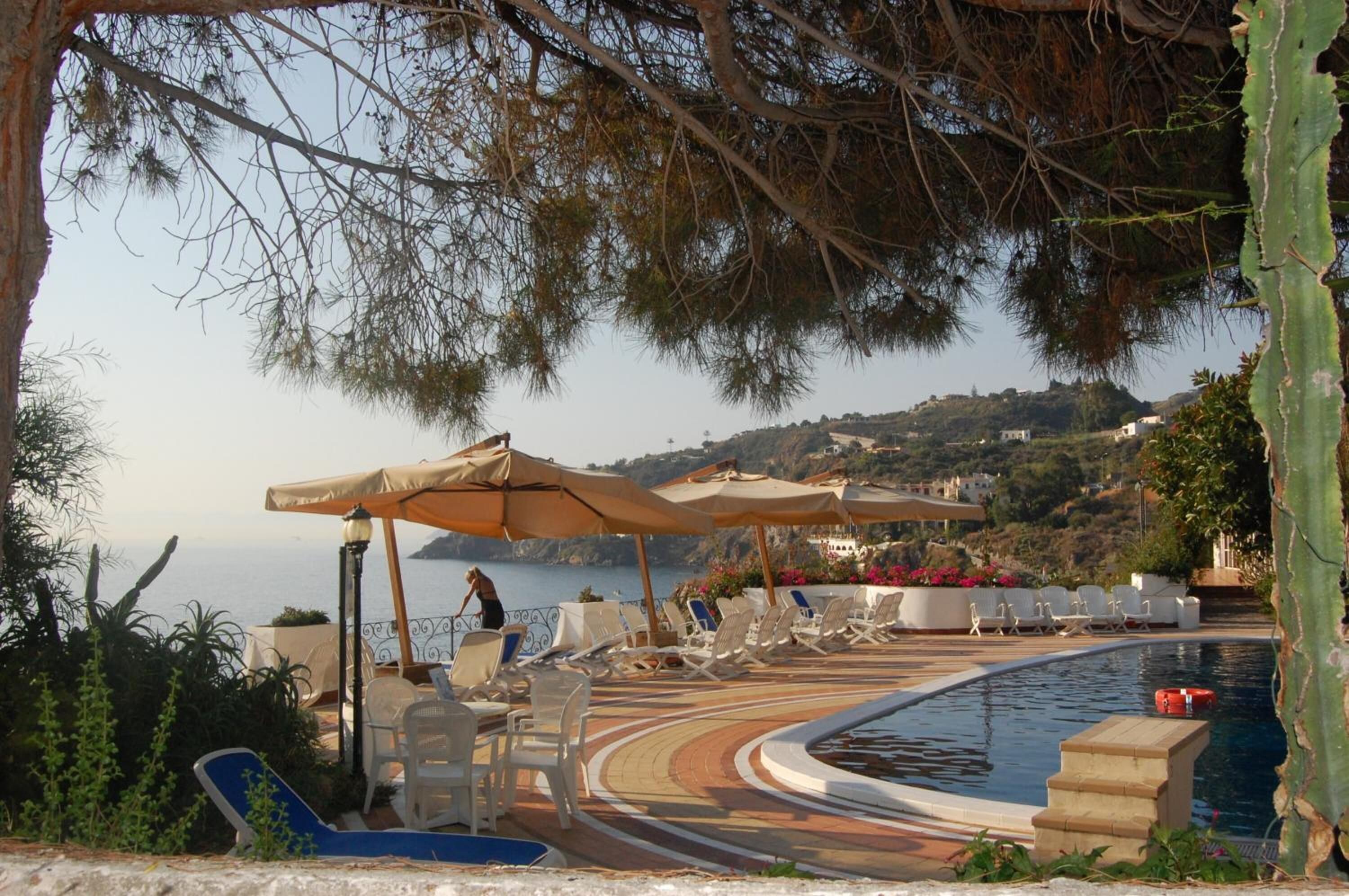 A poolside area with lounge chairs and umbrellas, overlooking a body of water and a hilly landscape.