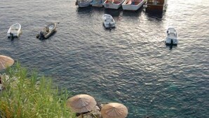 Una terraza frente al agua con varias embarcaciones amarradas al fondo, una hilera de sombrillas y un camino de piedra que lleva al agua.