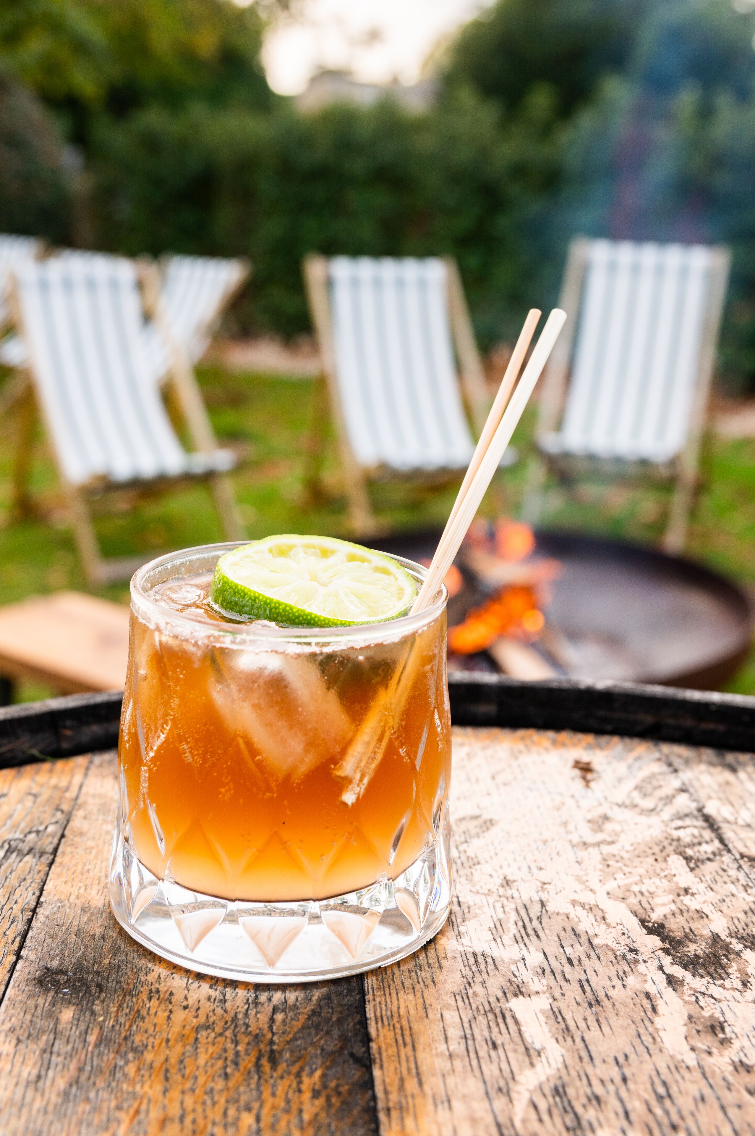 A glass of iced tea with a lime slice on a wooden table, with white striped chairs and a fire pit in the background.