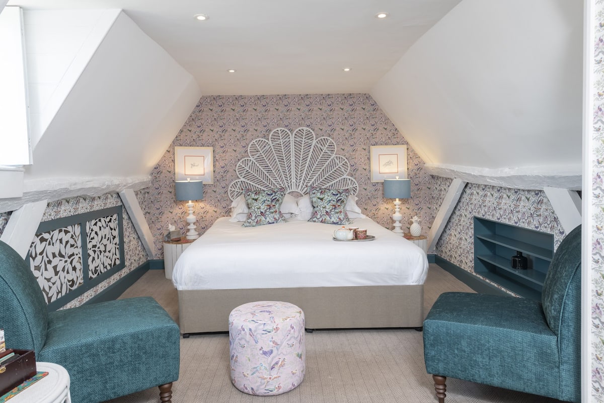 Quarto com cama grande, duas cadeiras, um pequeno banquinho, cabeceira decorativa e lareira.